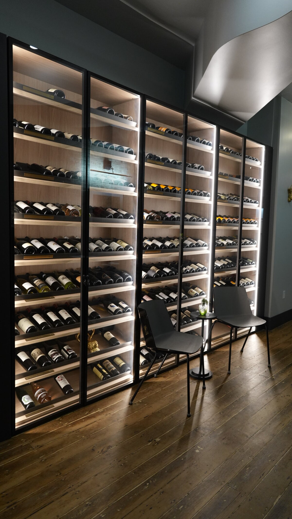 Close-up of red wine being poured into a glass at an elegant dinner setting, with a hand resting near a plate of gourmet food in a warmly lit restaurant.