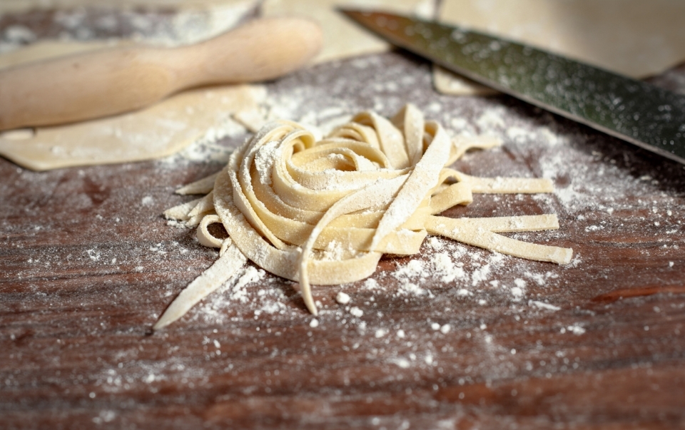 Freshly made fettuccine pasta nest on a floured wooden surface with a knife and rolling pin nearby.