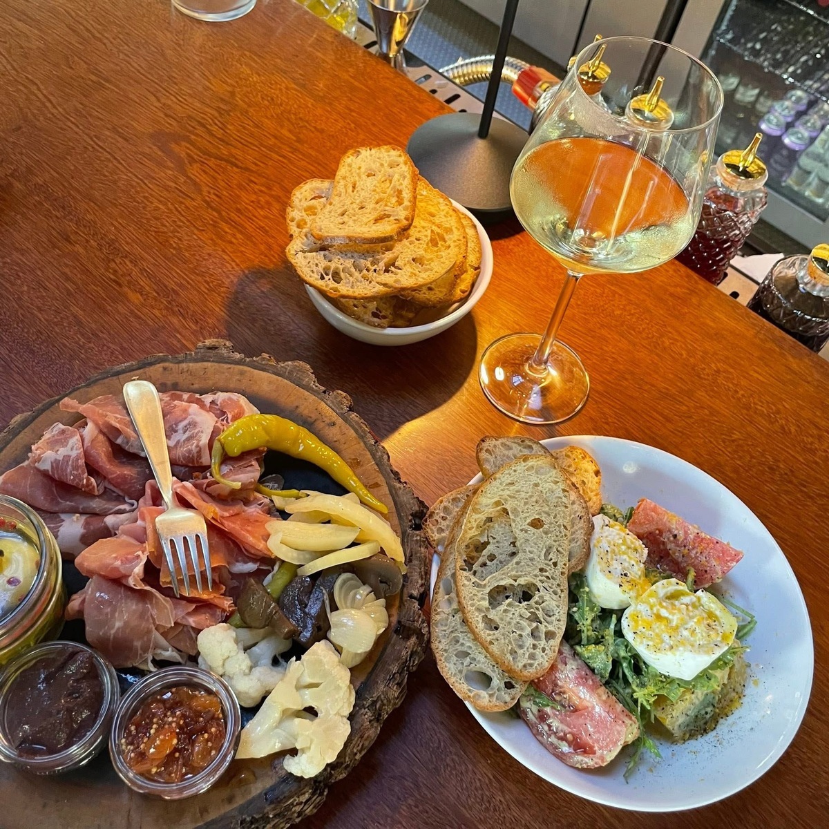 two full plates of food sitting on a wood tabletop next to a side of fresh bread and a glass of white wine